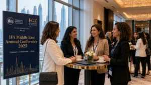 Four professional women standing around a high-top table, smiling and conversing at a conference. They each have conference lanyards and cups of coffee. A sign next to them reads “IFA Middle East Annual Conference 2025 – Connect | Collaborate | Grow,” with a skyline illustration. Other attendees are visible in the background.